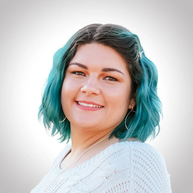 A woman with shoulder-length teal and black hair smiling at the camera, wearing a white knit sweater and hoop earrings