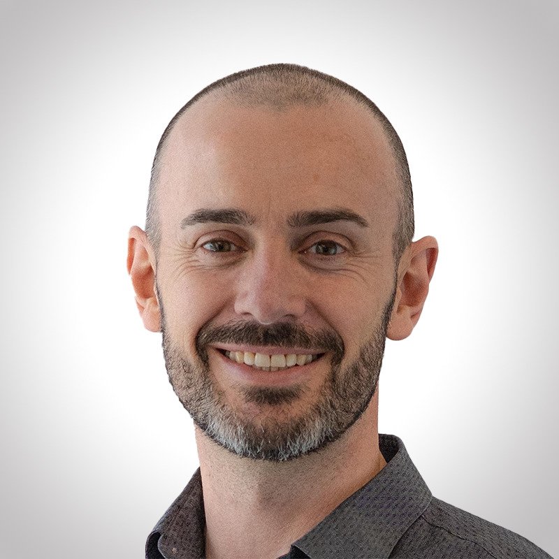 A smiling man with a shaved head and beard, wearing a dark collared shirt, against a plain white background.