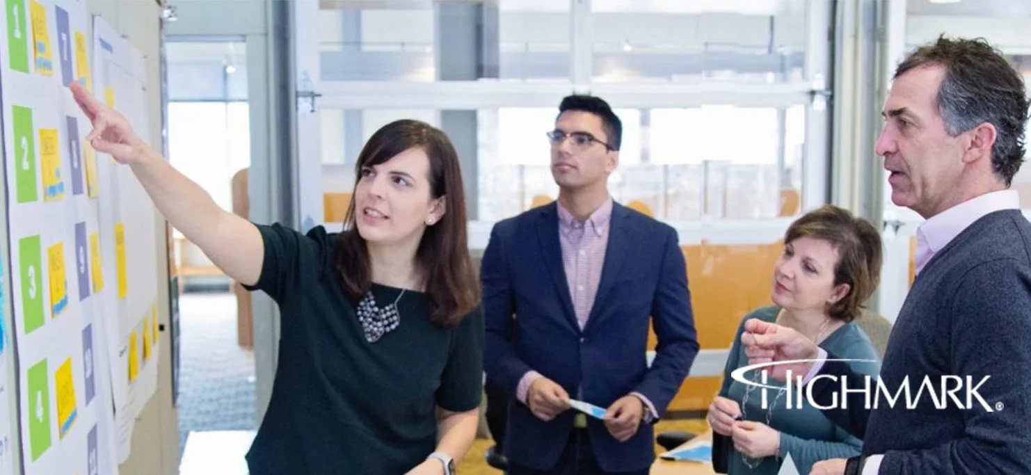 A woman explains something on a presentation board to a group of four people in a bright office setting, branded Highmark.