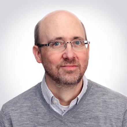 A middle-aged man with glasses, a beard, and a receding hairline wearing a gray sweater and collared shirt, against a plain light background.