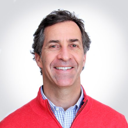 A smiling man with gray hair wearing a red jacket over a blue collared shirt, against a plain light background.