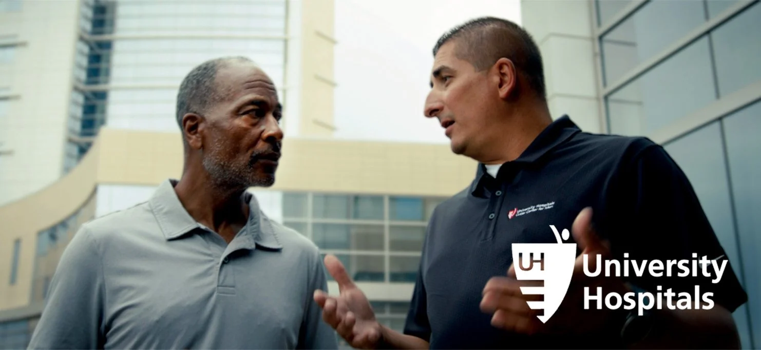 Two men having a conversation outside a building with modern architecture. One man is gesturing with his hands, branded University Hospitals.