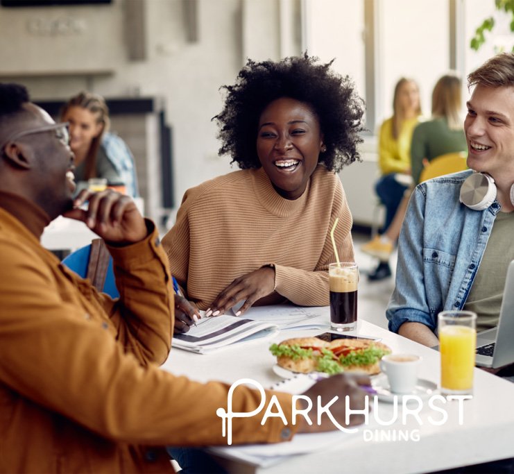 Three people sitting at a table in a lively restaurant, laughing and talking, with food and drinks on the table, with a woman with curly hair and a tan sweater in the center, branded Parkhurst.