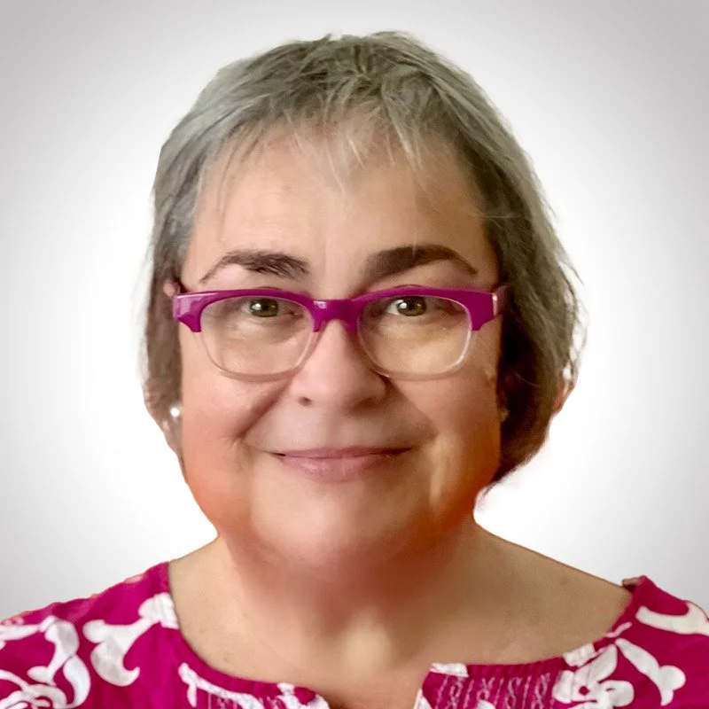 A middle-aged woman with short gray hair, pink glasses, and a pink and white patterned top, smiling against a plain light background.