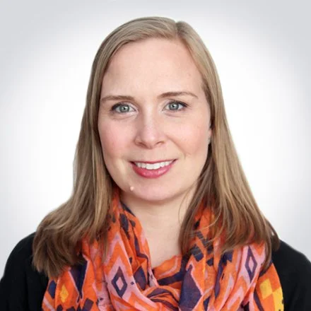 A woman with blonde hair wearing a black top and a colorful orange, pink, and purple scarf, smiling against a plain white background.