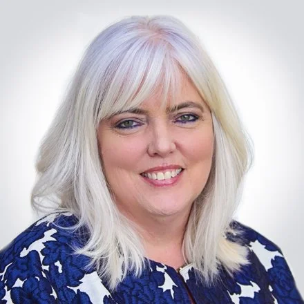 A woman with shoulder-length platinum blonde hair, smiling, wearing a blue and white patterned blouse.