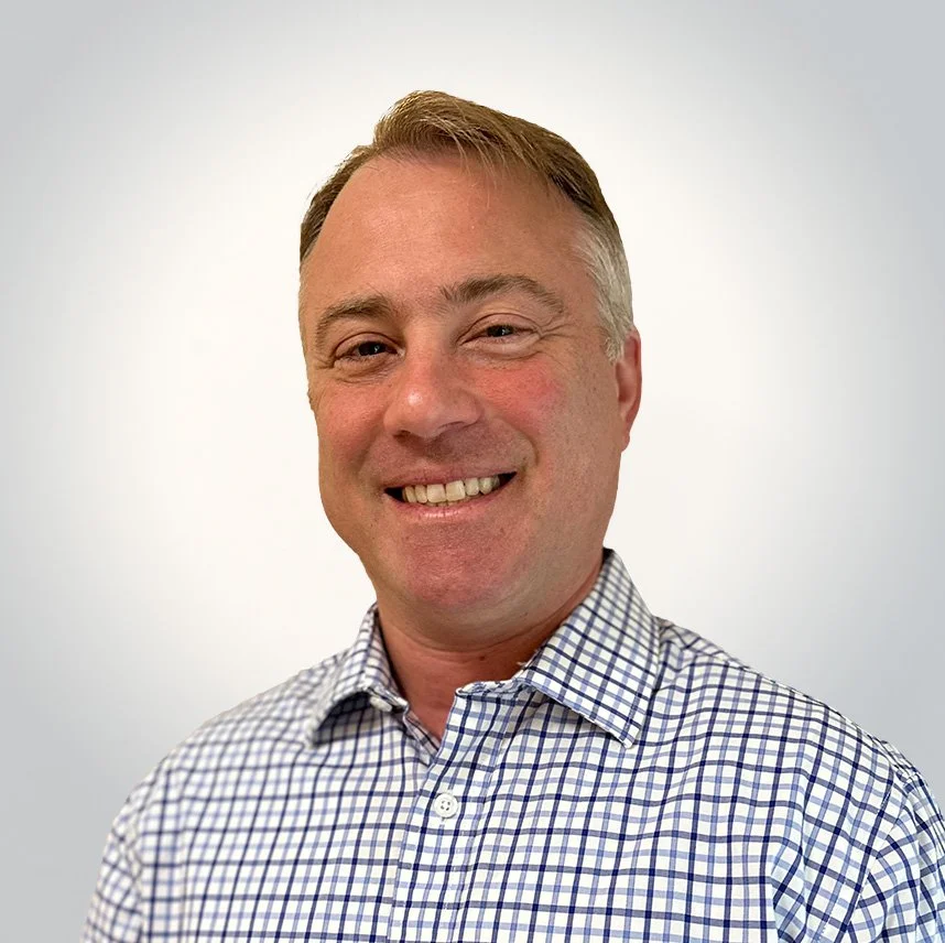 A smiling man with short blonde hair wearing a checkered dress shirt against a plain white background.