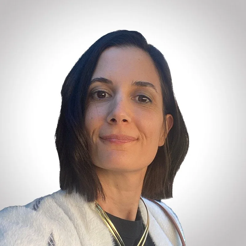 A woman with shoulder-length dark hair, wearing a light-colored sweater, a black top, and layered necklaces, smiling in front of a plain, light background.