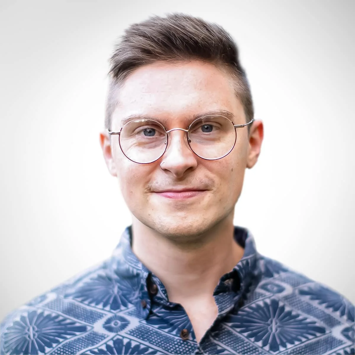 A young man with short brown hair, wearing round glasses, and a patterned button-up shirt with blue and black designs, standing against a plain white background.