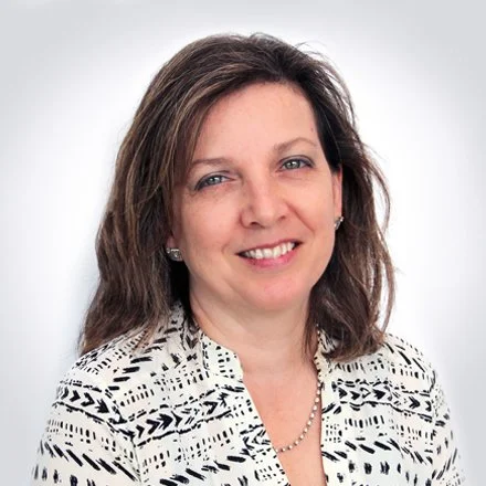 A woman with shoulder-length brown hair smiling, wearing a black and white patterned blazer, pearl earrings, and a pearl necklace against a plain gray background.