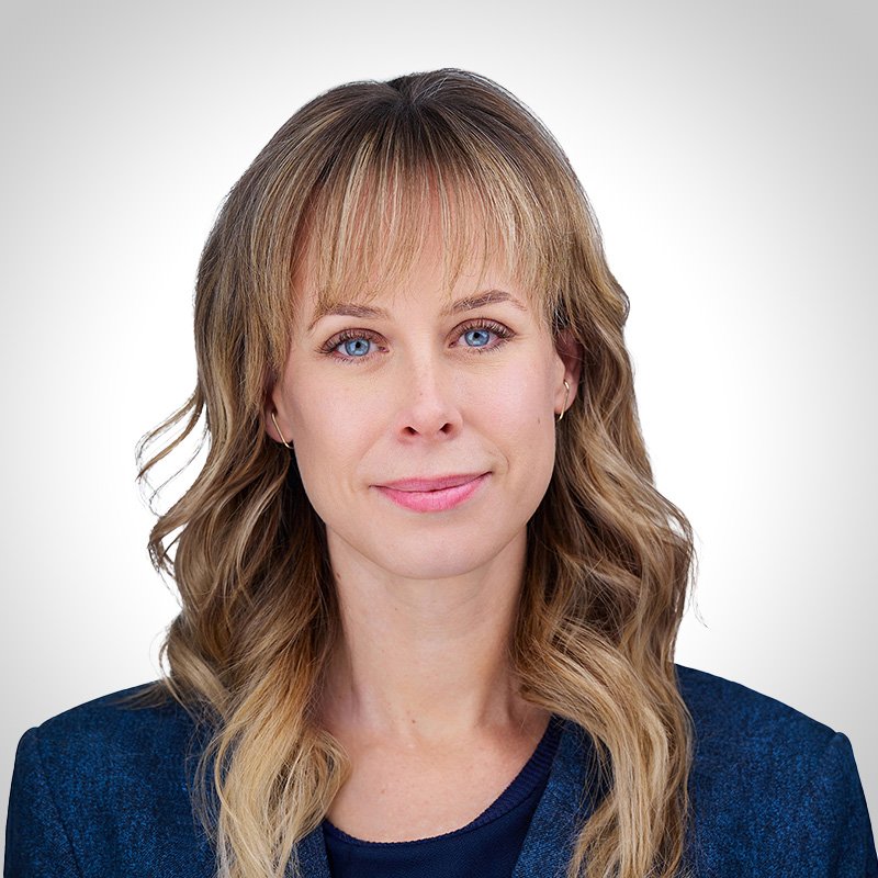 A woman with blonde, wavy hair and blue eyes, wearing a dark blue blazer and a navy top, looking at the camera with a neutral expression.
