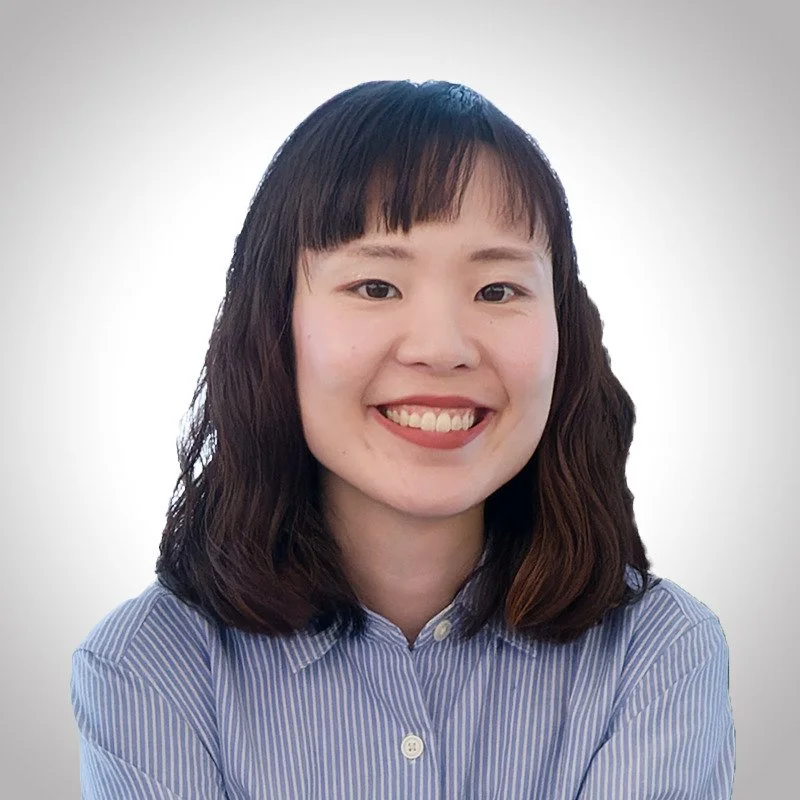 A young woman with shoulder-length dark hair and bangs, smiling, wearing a blue and white striped button-up shirt, against a light gray background.