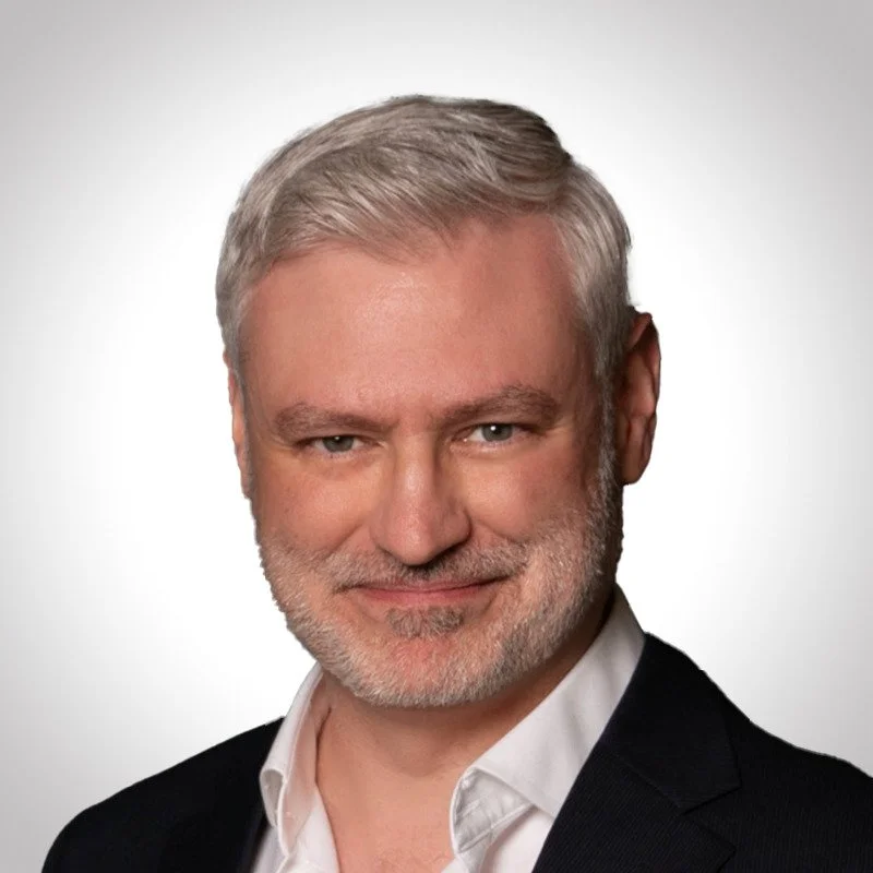 A middle-aged man with gray hair and beard, wearing a black blazer and white shirt, smiling slightly against a plain light background.