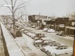 Black and white vintage photo of a busy street with parked cars and small retail shops.