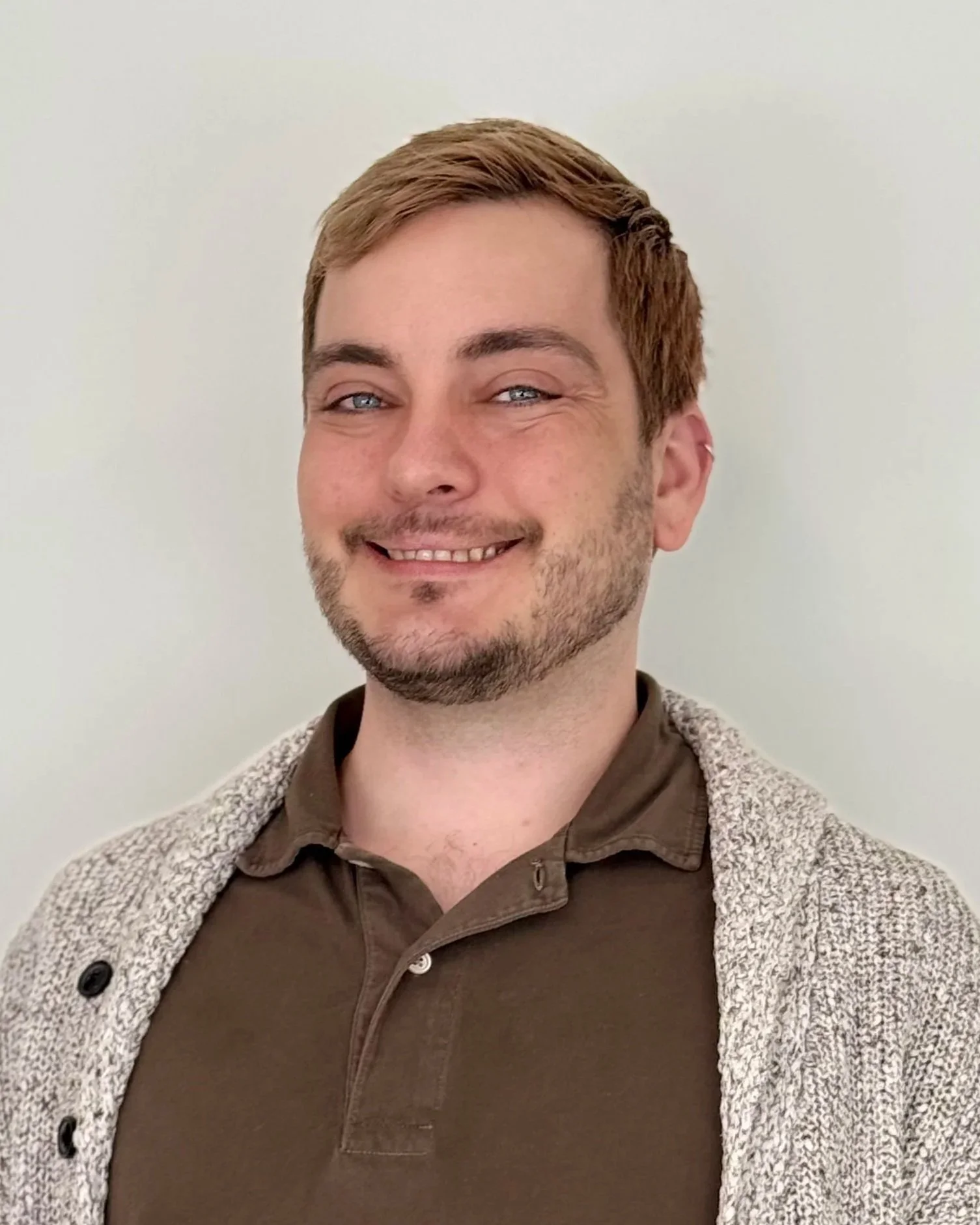A smiling man with light brown hair and blue eyes, wearing a brown shirt and a gray cardigan, standing against a plain white wall.
