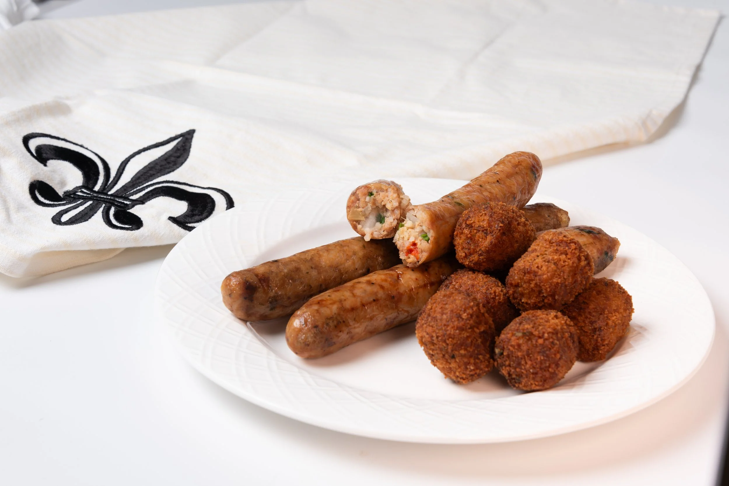 Plate with cooked boudin and breaded boudin balls, with a white cloth featuring a black fleur-de-lis in the background.