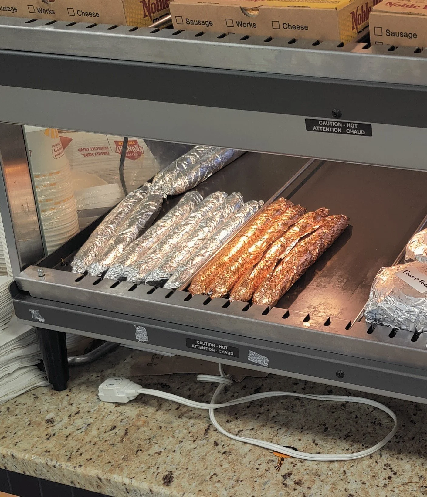 Boudin links wrapped in foil on a heating tray inside a food warmer, with a caution sign indicating heat.