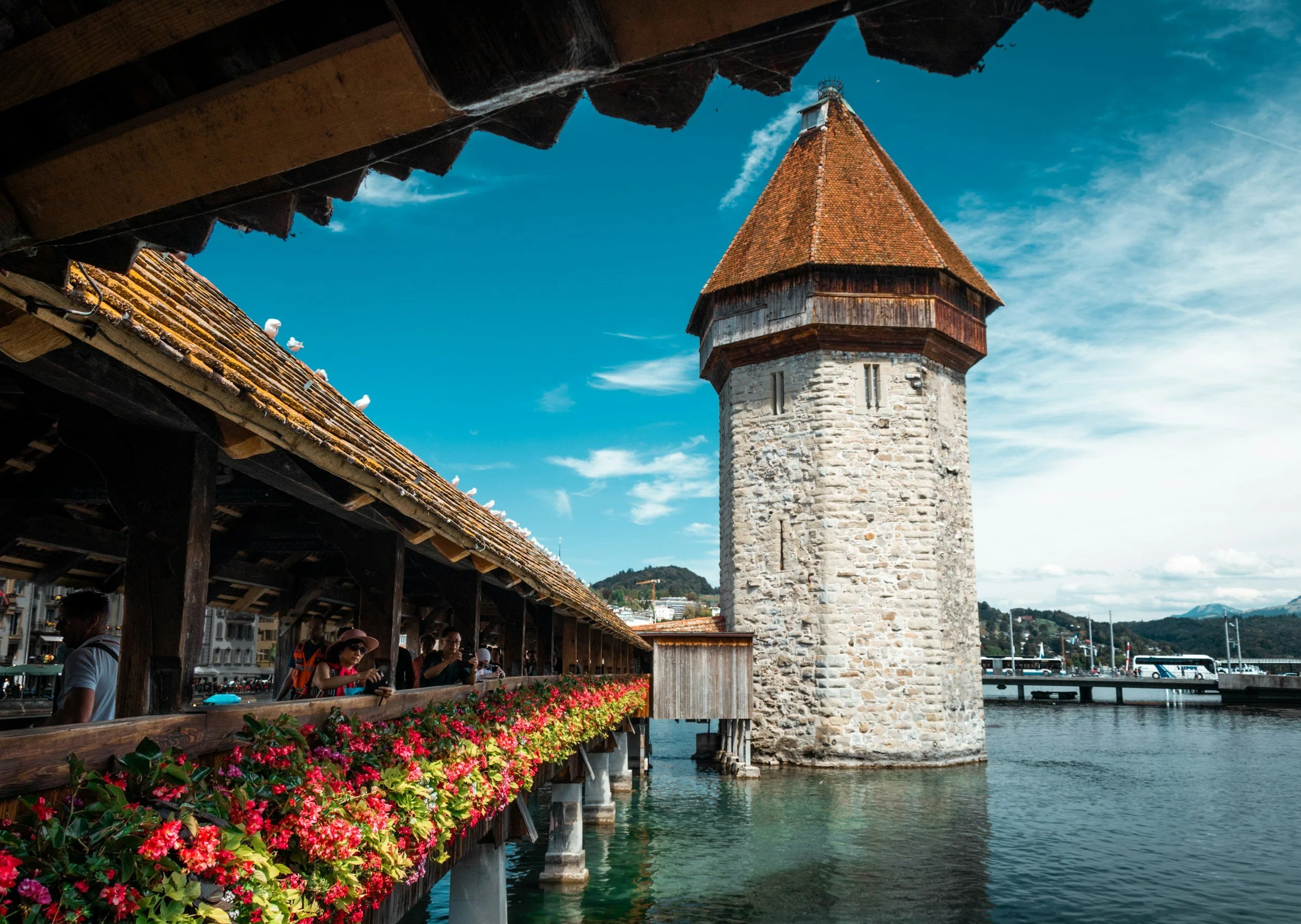 Historischer Turm neben einer alten Holzbrücke mit Blumenschmuck und Touristen bei schönem Wetter in einer Stadt am Wasser.