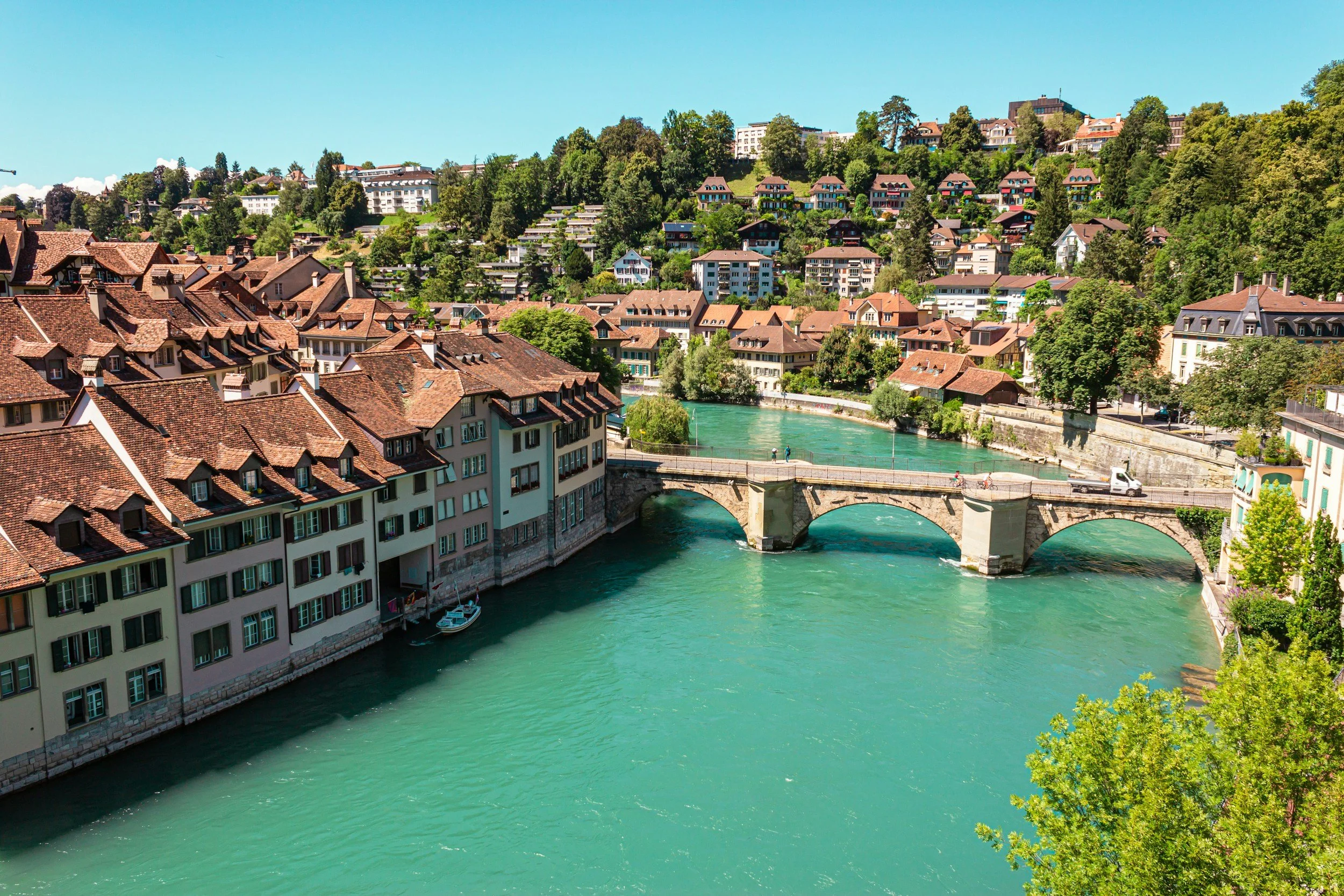 Blick auf einen Fluss, der durch eine Stadt in die Berge fließt, mit einer Brücke, traditionellen Häusern mit roten Dächern und grüner Landschaft im Hintergrund.