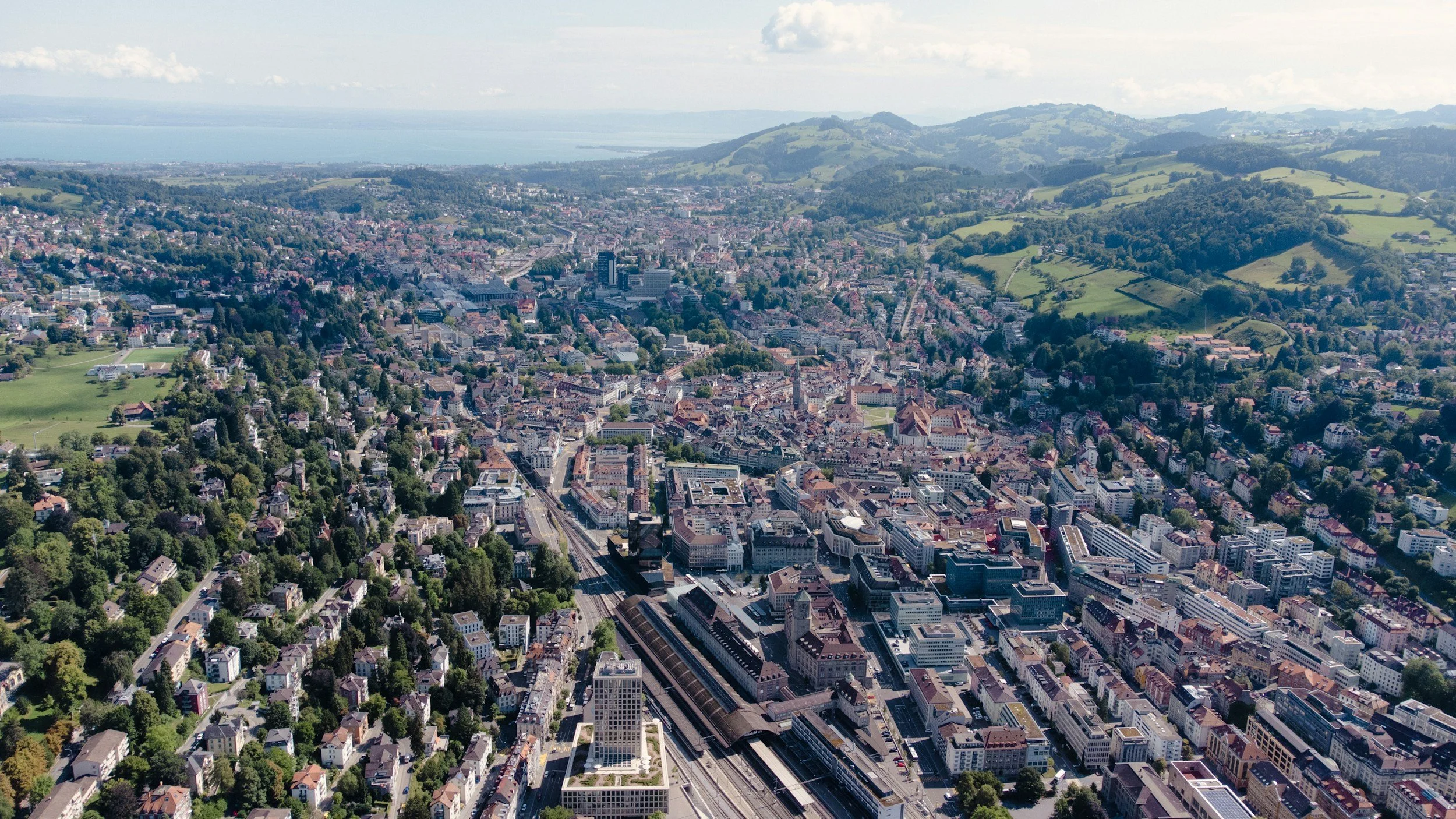 Vogelperspektive auf die Stadt Zürich mit Bahnhof, umliegenden Gebäuden und grünen Hügeln im Hintergrund.