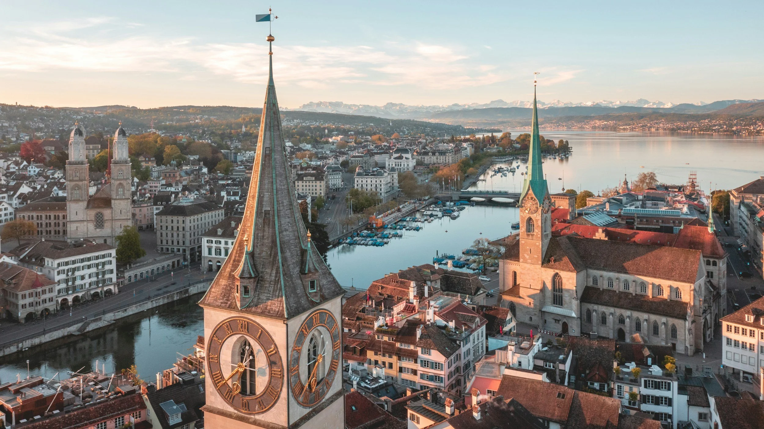 Luftaufnahme einer Stadt mit zwei Kirchtürmen, einem Fluss und Bergen im Hintergrund bei Sonnenuntergang.