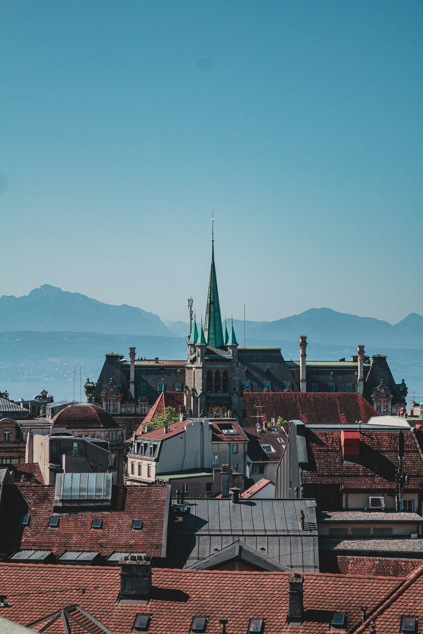 Blick auf eine Stadt mit roten Dächern und einem großen alten Gebäude mit einem hohen Kirchturm im Hintergrund, umgeben von Bergen