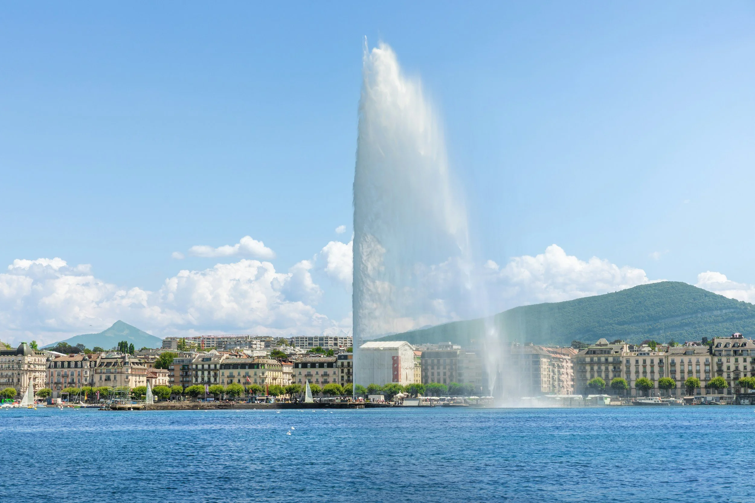 Der Blick auf den Genfersee mit dem berühmten Wasserstrahllauf in der Stadt Genève, Schweiz, mit Gebäuden am Ufer, grünen Bäumen und Hügeln im Hintergrund bei sonnigem Wetter.