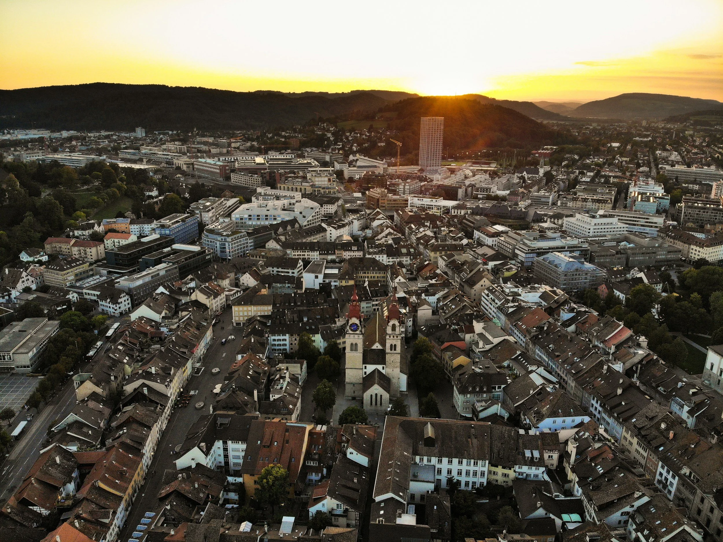 Luftaufnahme einer Stadt bei Sonnenuntergang, mit historischen und modernen Gebäuden, umgeben von Hügeln.