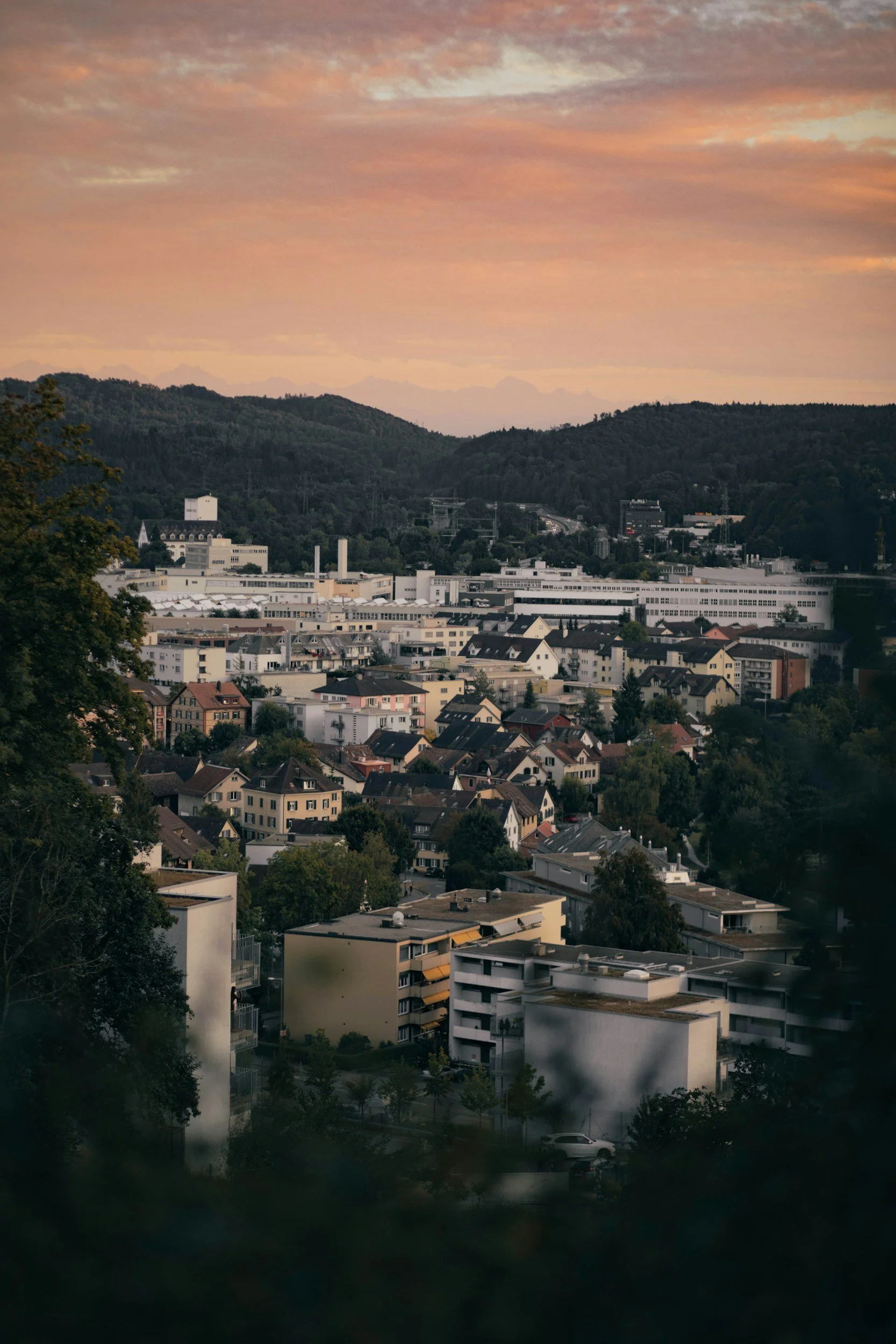 Stadtansicht bei Sonnenuntergang mit Häusern, Bäumen und Hügeln im Hintergrund.
