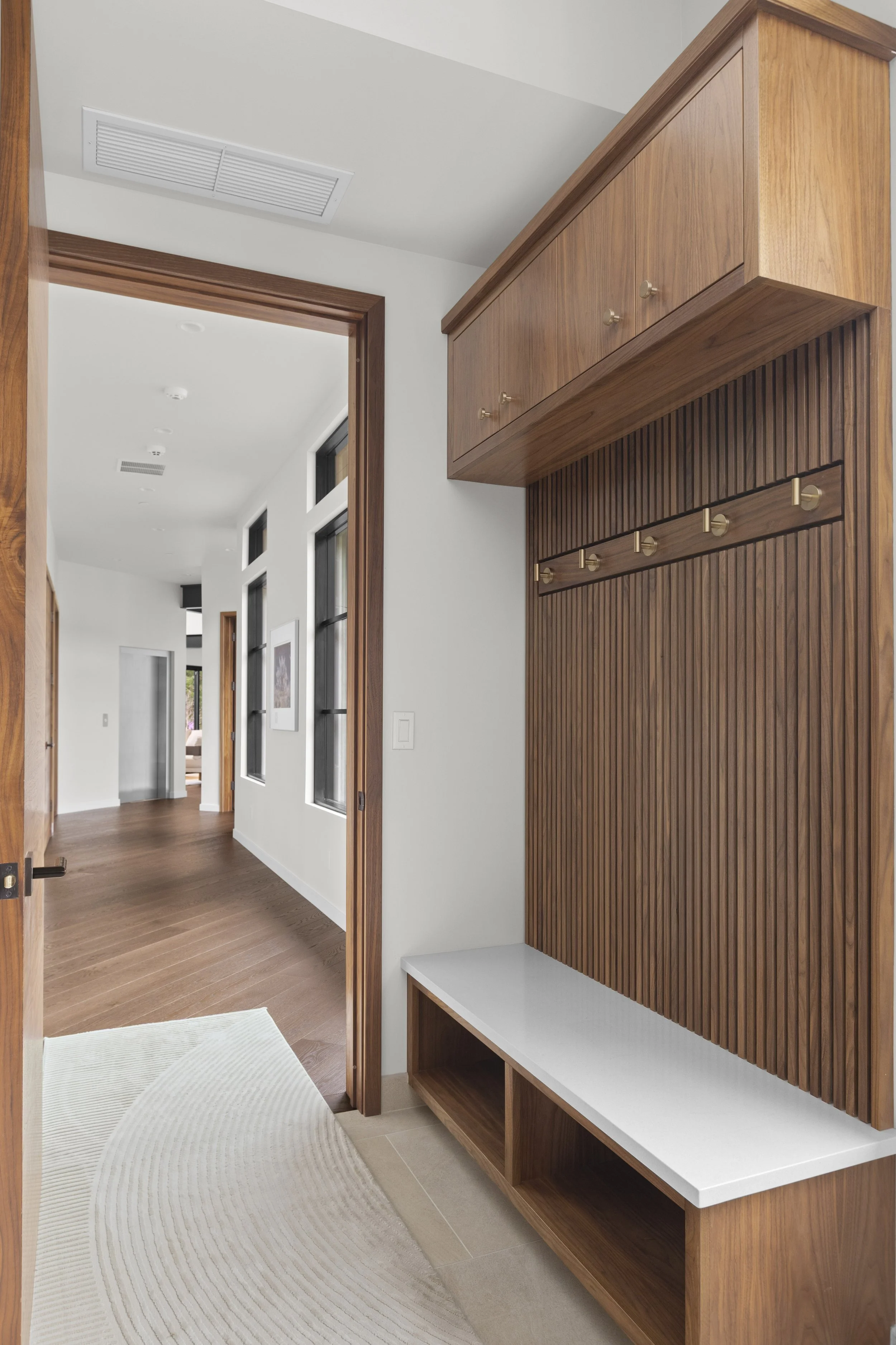 Custom walnut mud room bench with slatted panel wall, brass hooks, and overhead storage