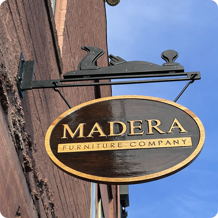 Madera Furniture Company shop sign: carved wood oval hanging from a blackened steel bracket with a hand plane silhouette, Tacoma.