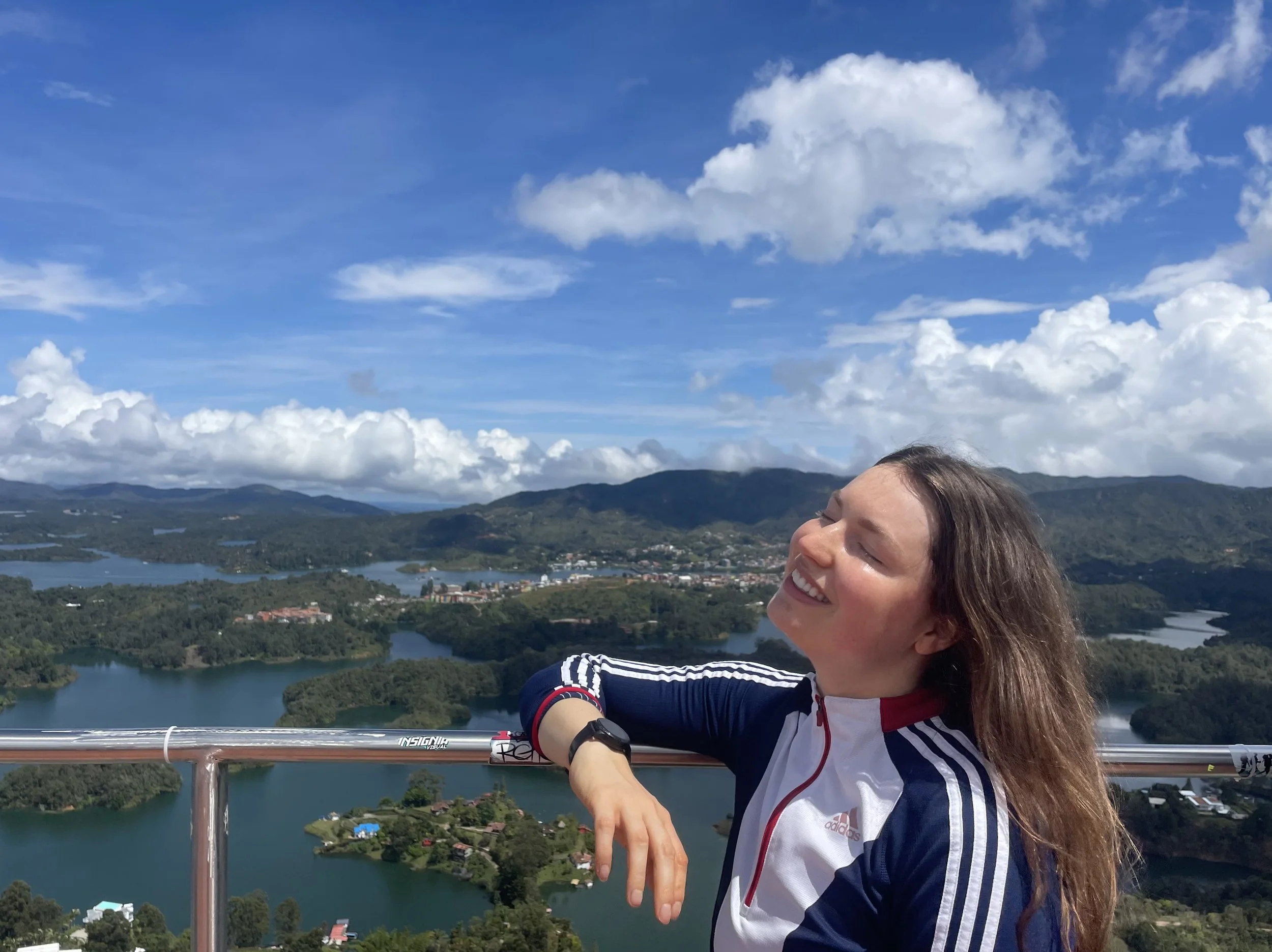 Mujer sonriendo con los ojos cerrados, disfrutando del paisaje desde una terraza, con un fondo de montañas, lagos y cielo con nubes.
