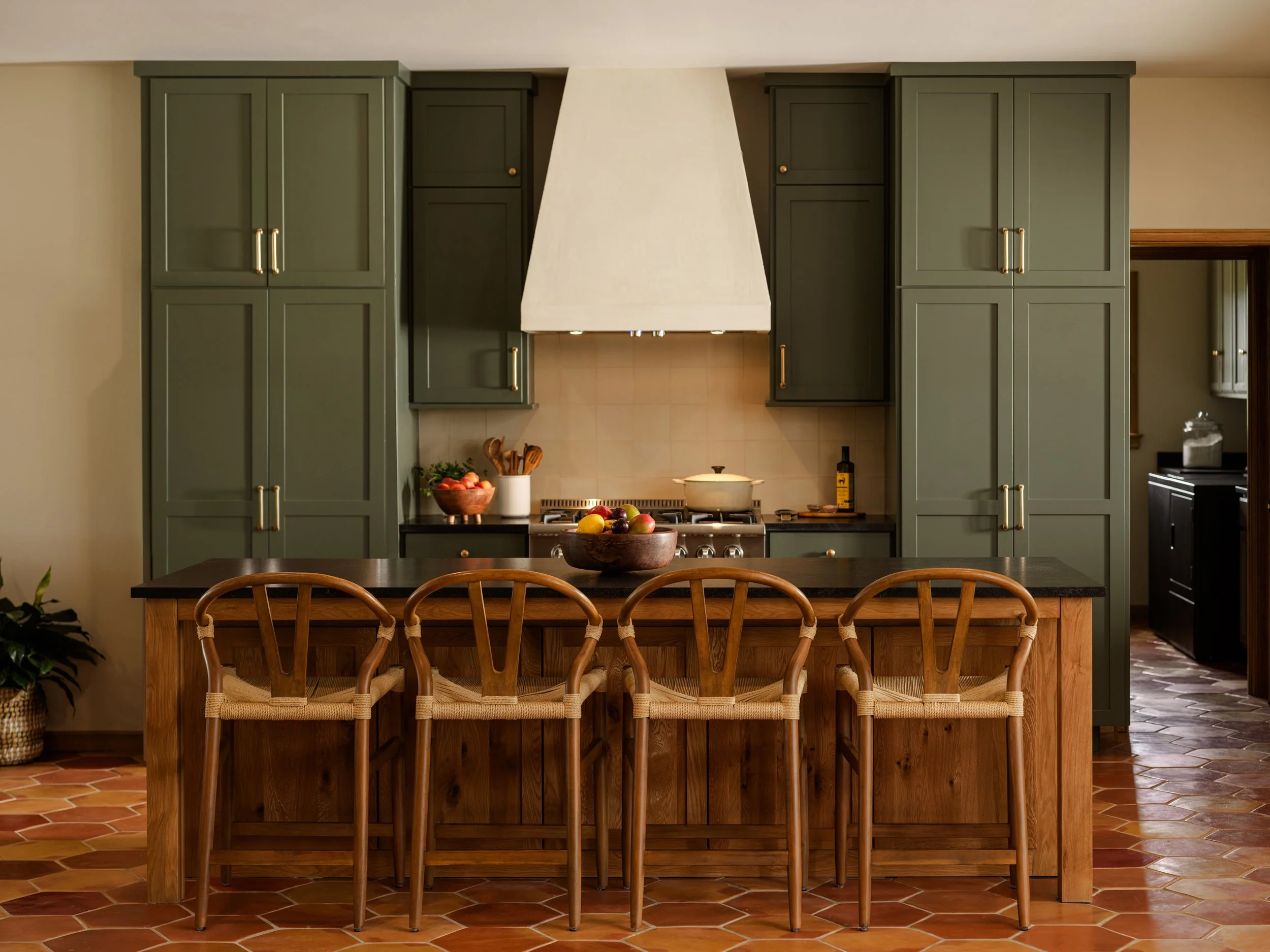 Kitchen with green cabinets, wooden island, and four wooden chairs, featuring a stove with a pot, and a bowl of fruit on the island. Parade of Homes in Fredericksburg, TX, Kerrville, Burnet, Marble Falls, Austin, San Antonio, New Braunfels