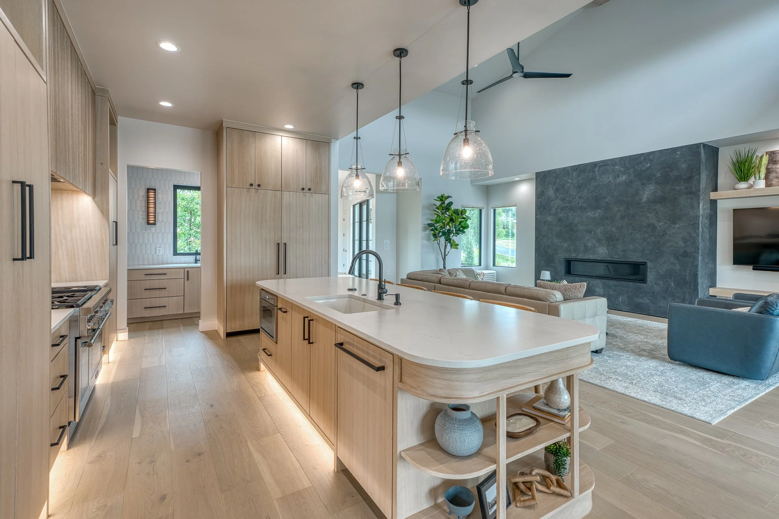 Open-concept kitchen and living room with light wood cabinets, an island with a sink, pendant lighting, a gray accent wall with a fireplace, and large windows with views of greenery.