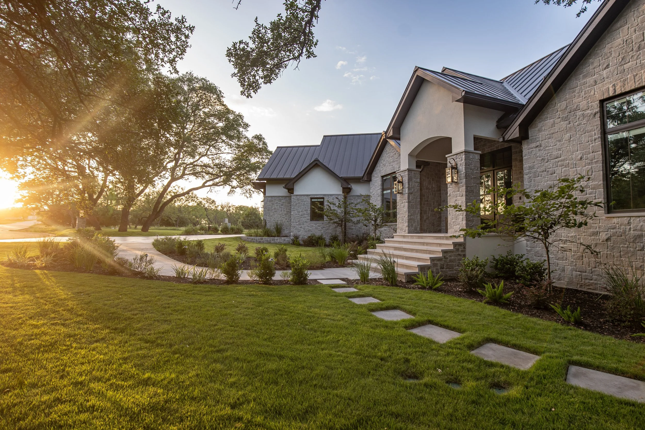Front view of a modern house with a stone exterior, staircase, large windows, and a black metal roof, surrounded by a landscaped yard and trees during sunset.