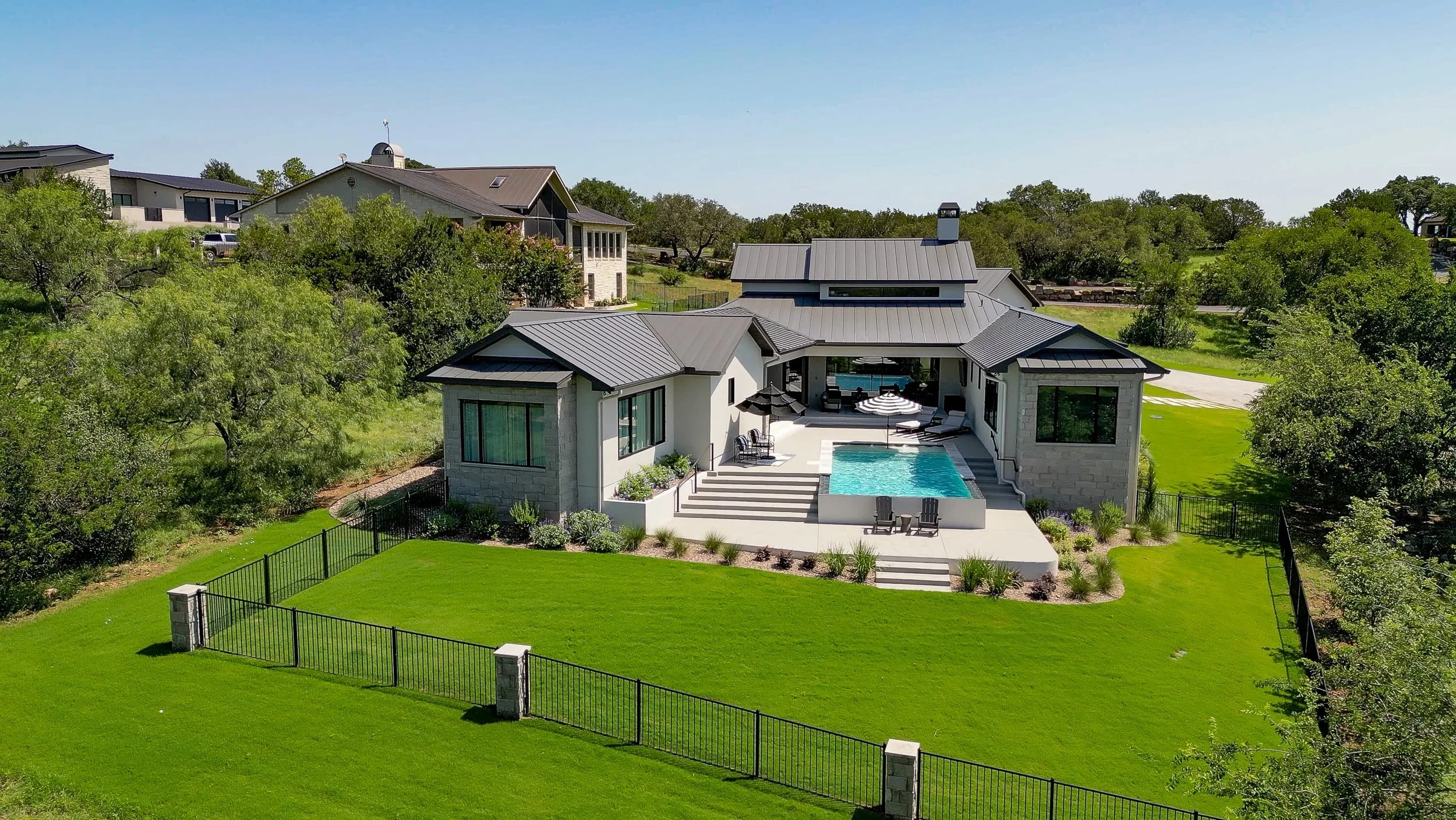 A modern house with a small rectangular swimming pool, outdoor furniture, and a well-maintained lawn surrounded by trees and a black fence.