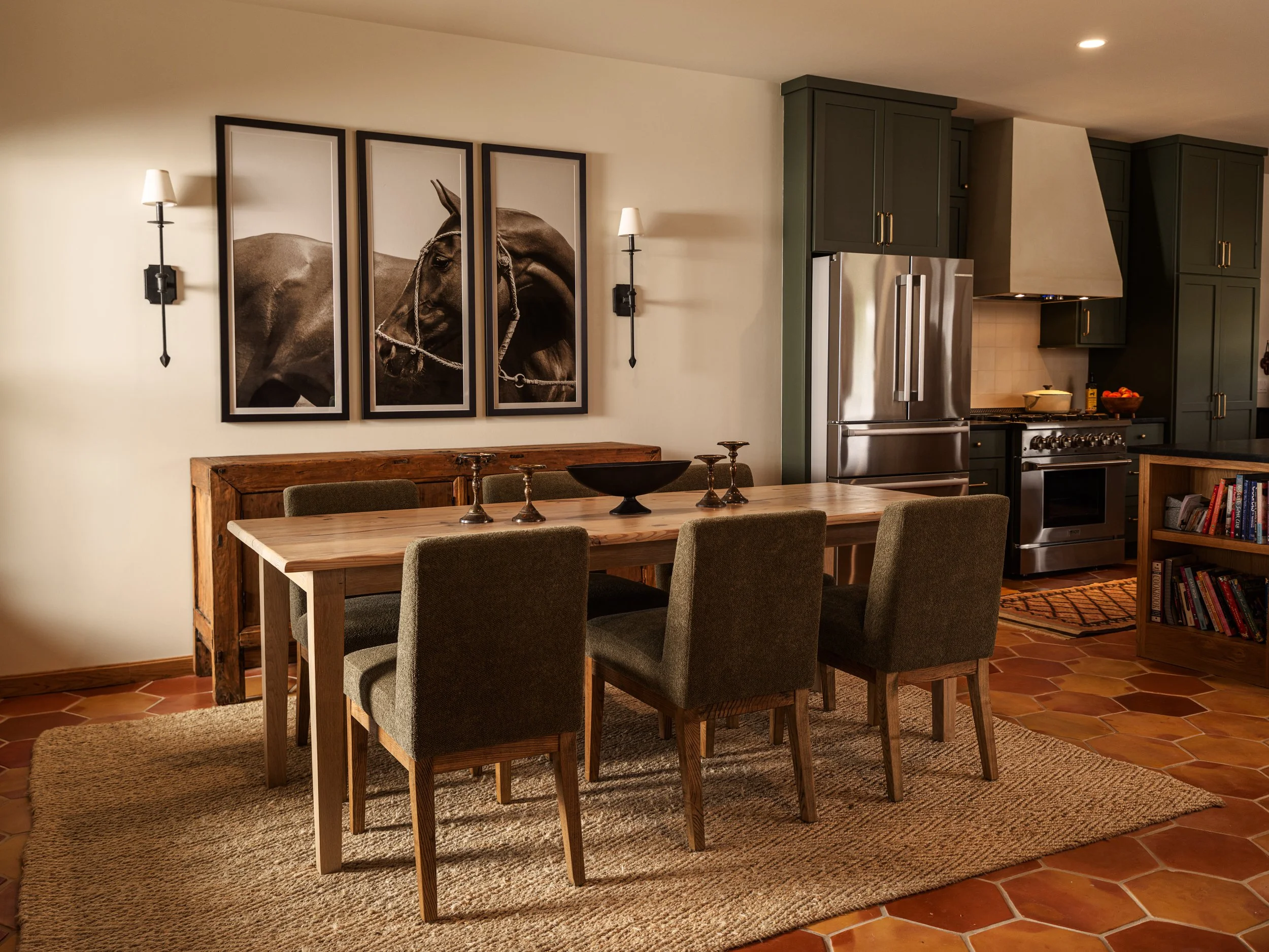 Dining room with a wooden table, six upholstered chairs, and a large black-and-white triptych of a horse on the wall. Kitchen area with stainless steel refrigerator, stove, green cabinets, and a bookshelf.