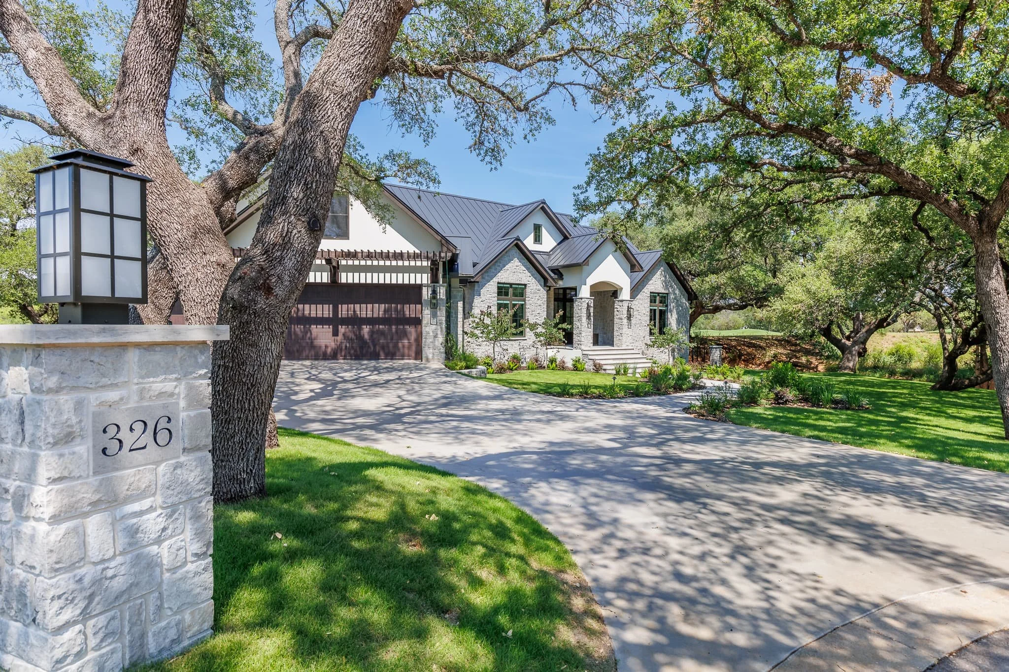 Front view of a modern house with a stone and white exterior, black roof, and a large driveway, surrounded by mature trees and green lawn.