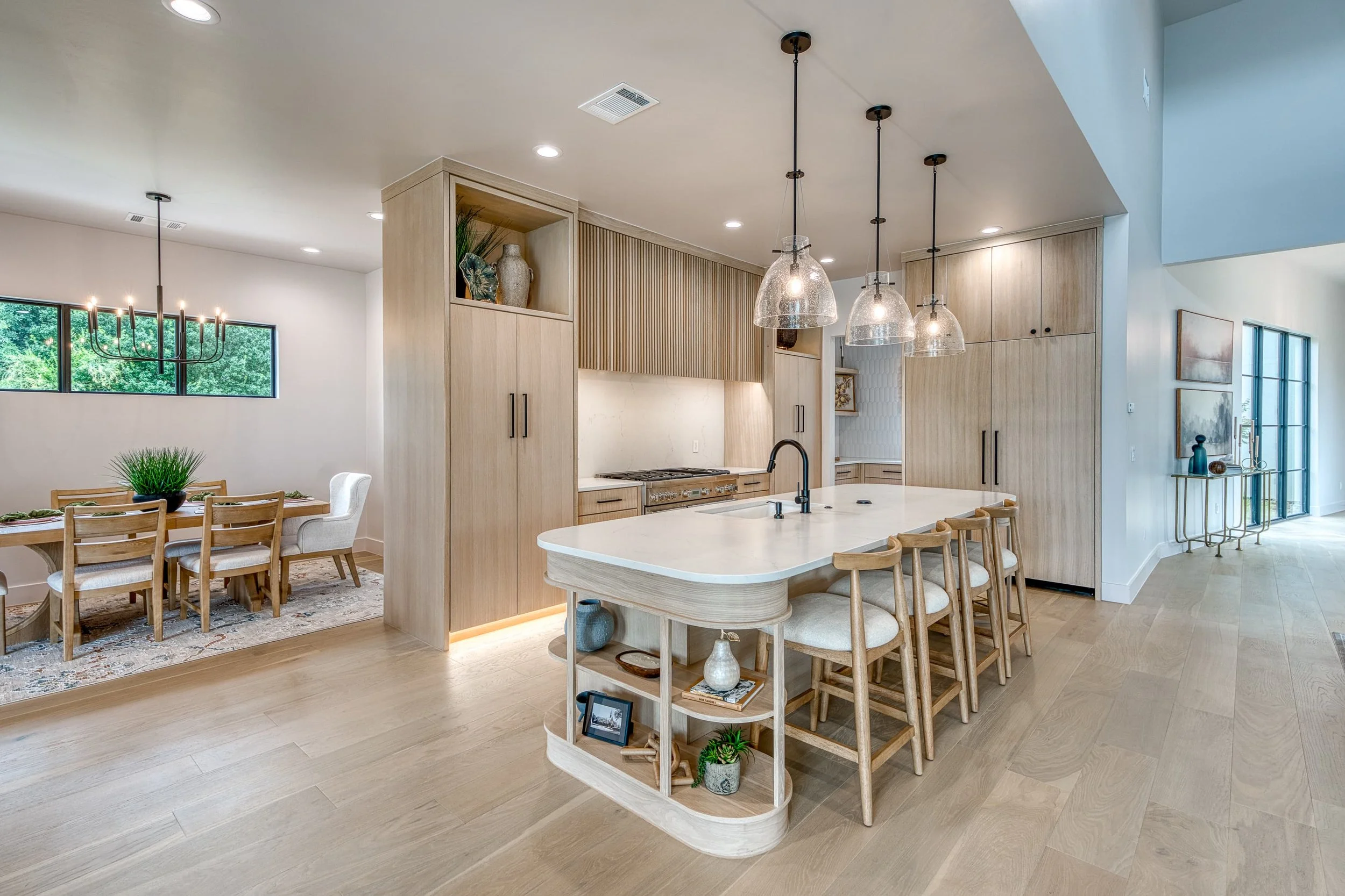 Open-concept kitchen and dining area with a light wooden island, beige cabinetry, pendant lights, and a dining table with chairs in a modern home.
