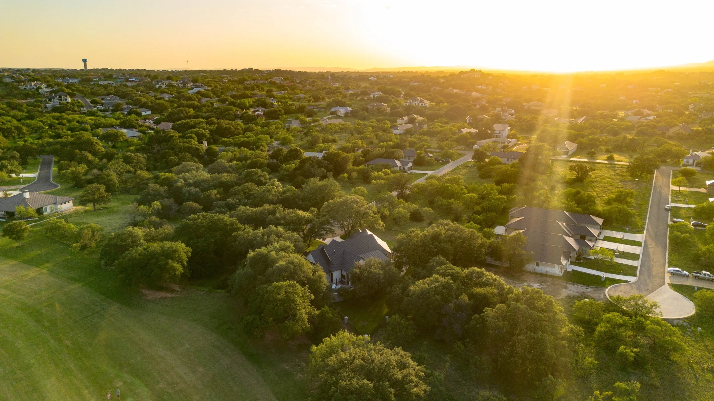 An aerial view of a suburban neighborhood at sunset with green trees, houses, and a large lawn. Fredericksburg TX custom home in the Hill COuntry Parade of Homes.