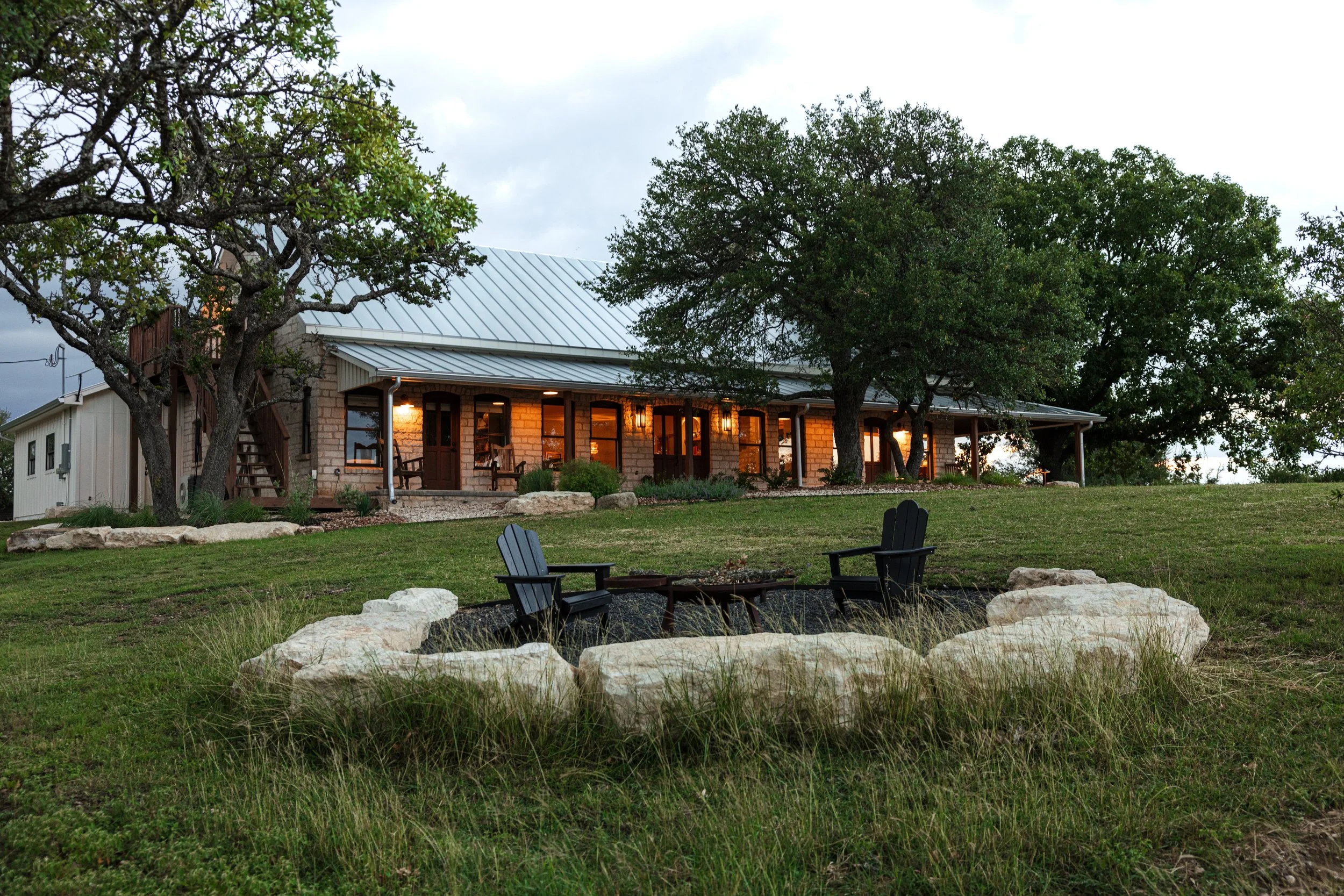 A house with a metal roof illuminated by warm outdoor lighting, surrounded by large trees and a grassy yard. There is a fire pit with two black chairs in the foreground.