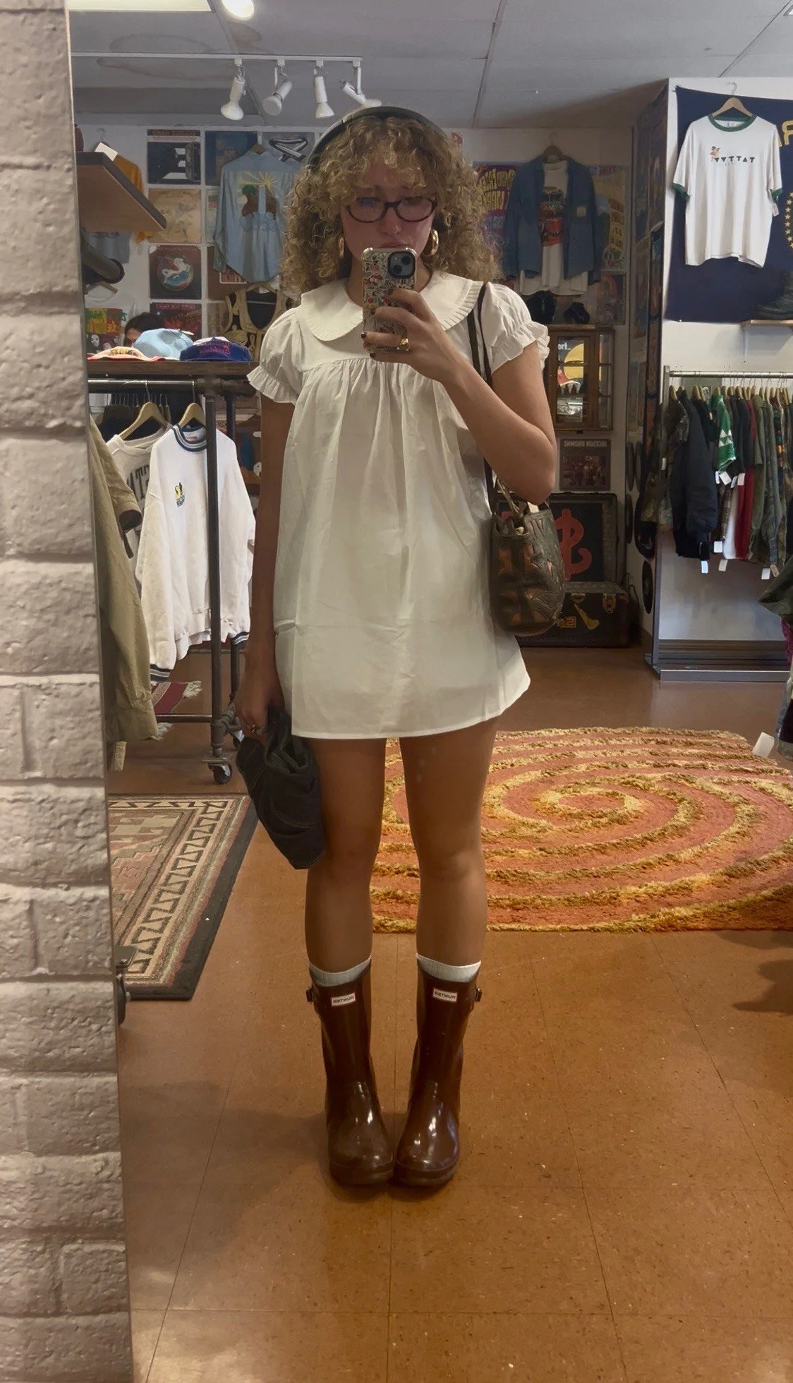 A woman with curly hair and glasses taking a mirror selfie in a vintage store. She is wearing a white dress, brown rubber rain boots, and white socks. She has a shoulder bag and is holding a black item in her hand.