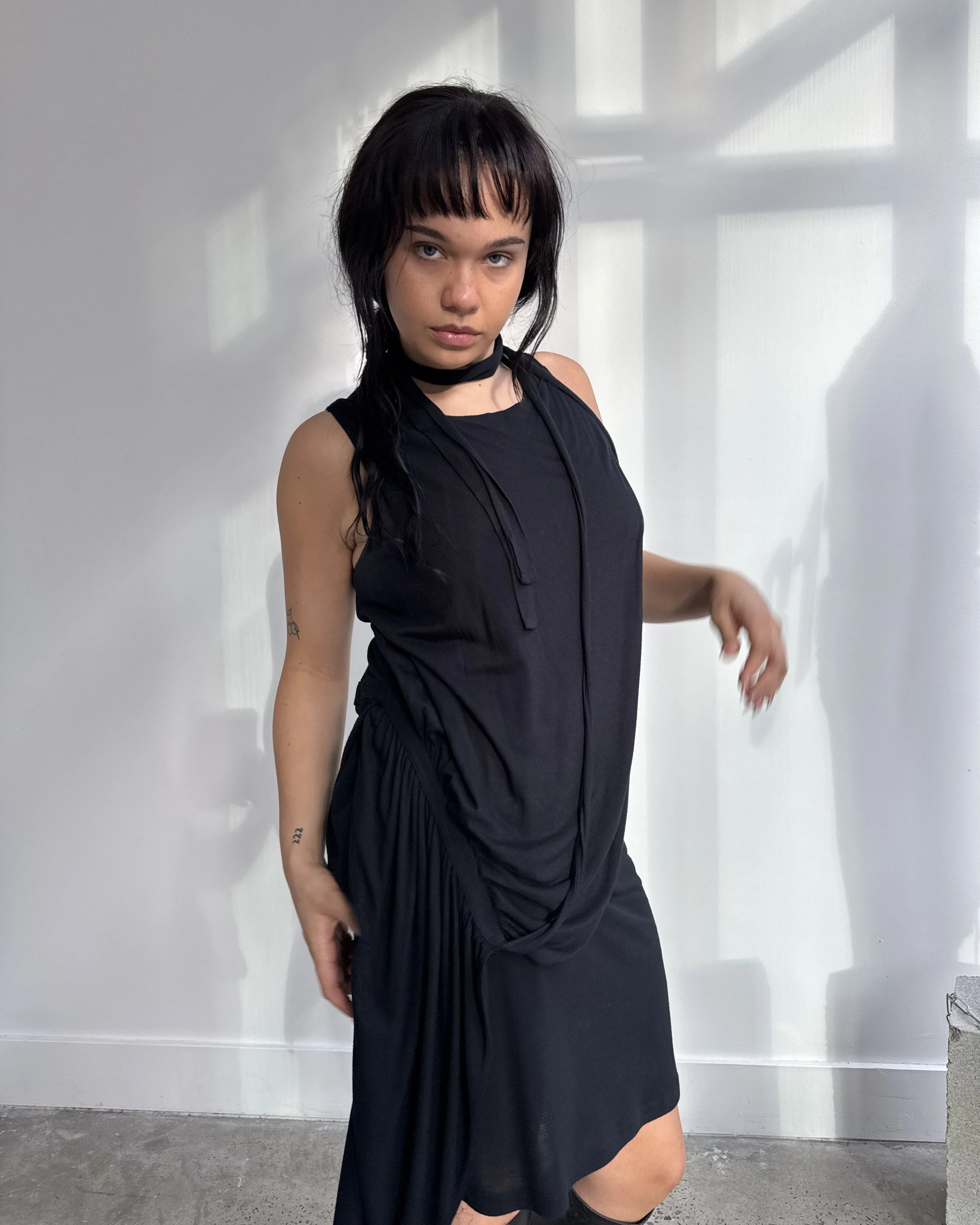 A young woman with black hair and bangs, wearing a sleeveless black dress, standing in front of a white wall with shadows of window frames.