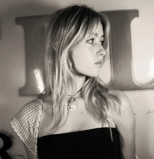 A young woman with wavy hair and a choker necklace, looking to the right, inside a vehicle with a window and curtain in the background, in black and white.