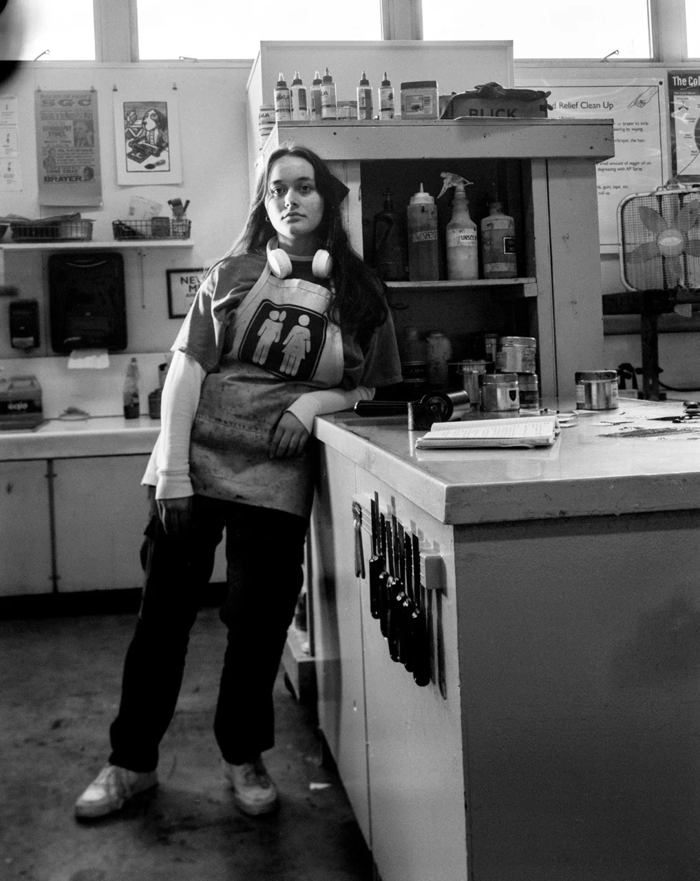 A young woman with long dark hair standing in a printshop, leaning against a counter with various printmakingtools and supplies. She is wearing an apron with a graphic of two figures and headphones around her neck.