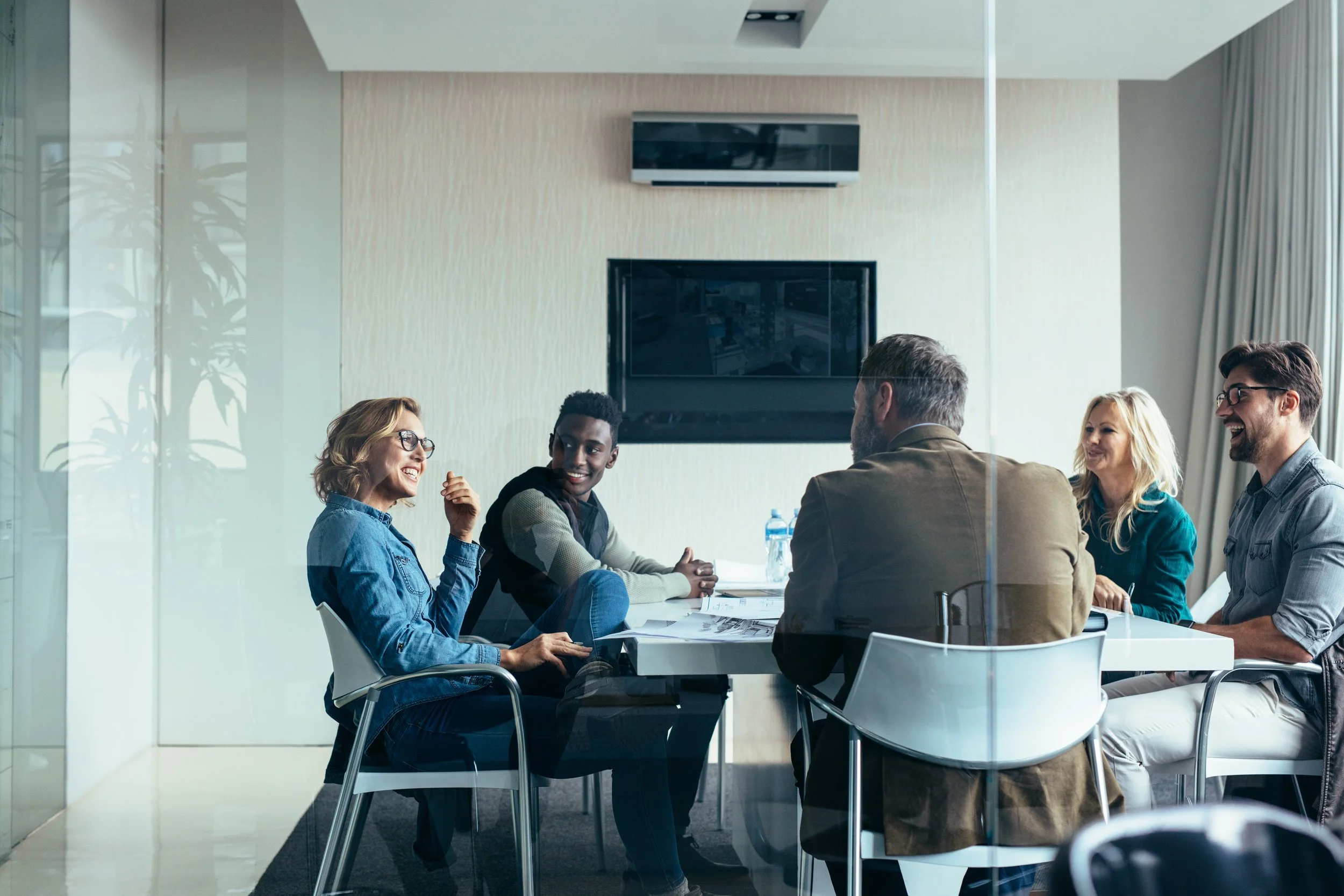 Five people sitting at a white conference table in a modern meeting room, engaged in conversation and smiling.