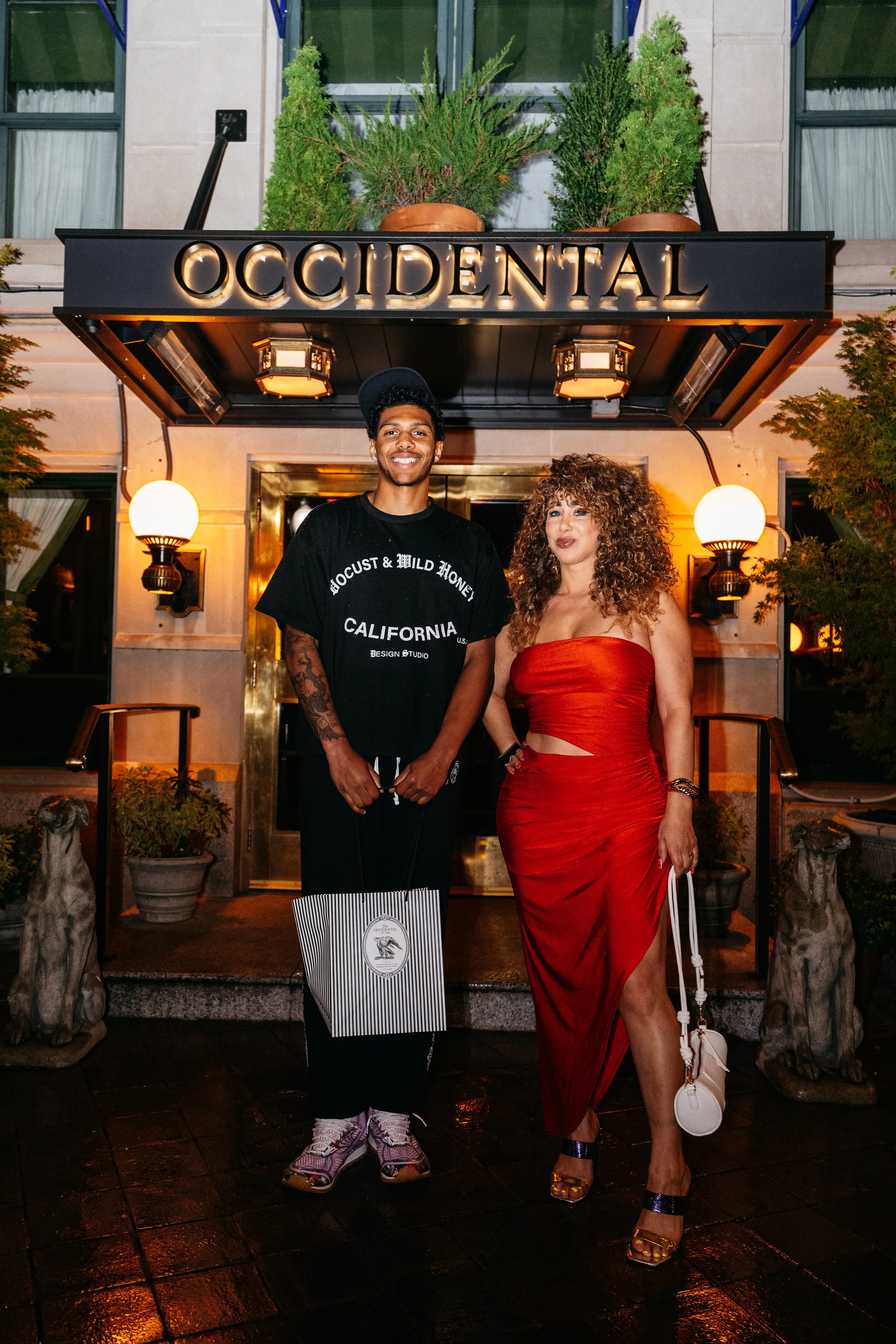 Two people standing in front of the Occidental hotel entrance, with a sign reading 'OCCIDENTAL' above them, and decorative plants and statues on either side.