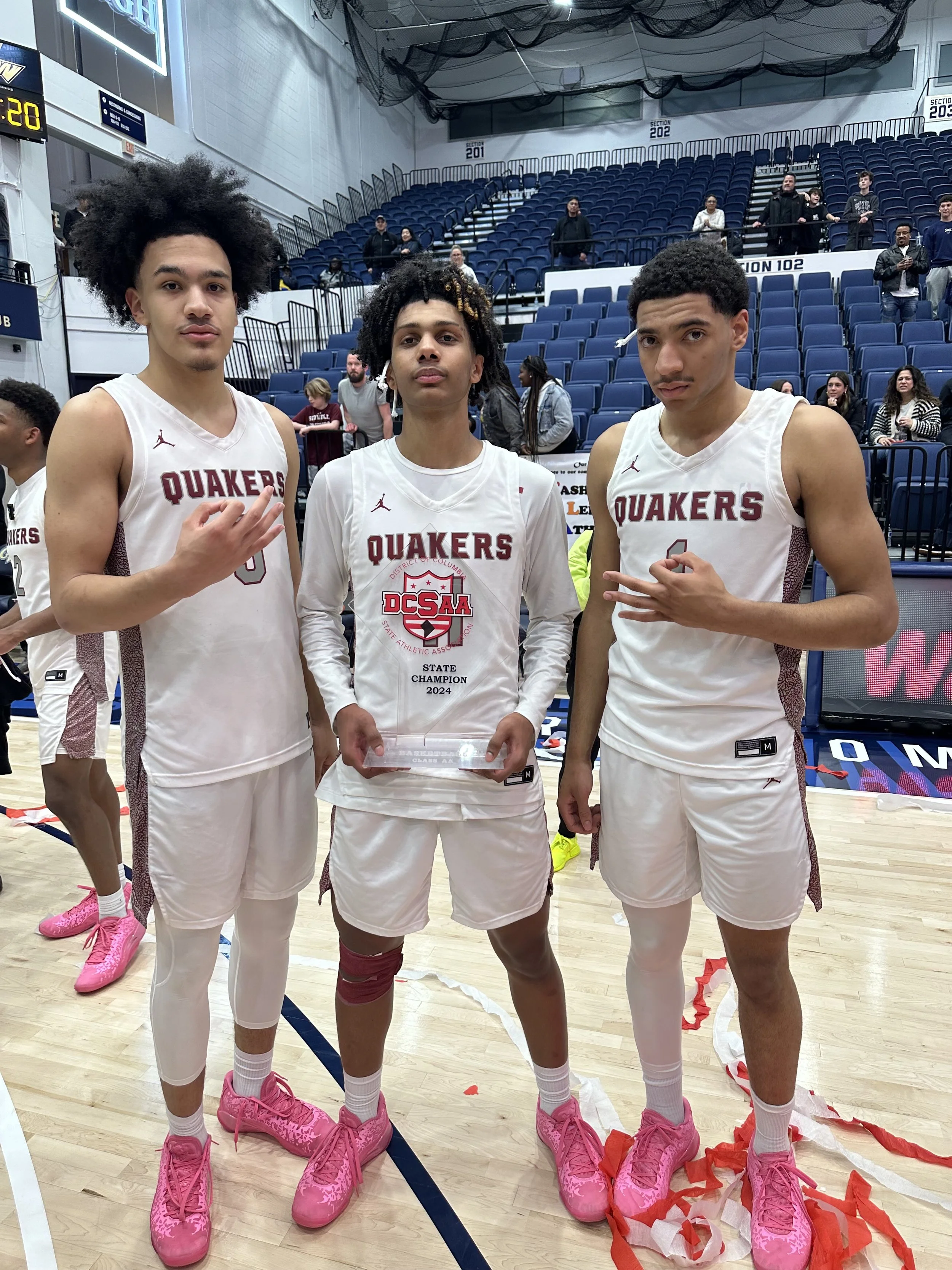 Three basketball players on a court celebrating their championship win. They are wearing white Quakers jerseys and pink shoes, with one holding a trophy. The gym has blue seating and a scoreboard in the background.