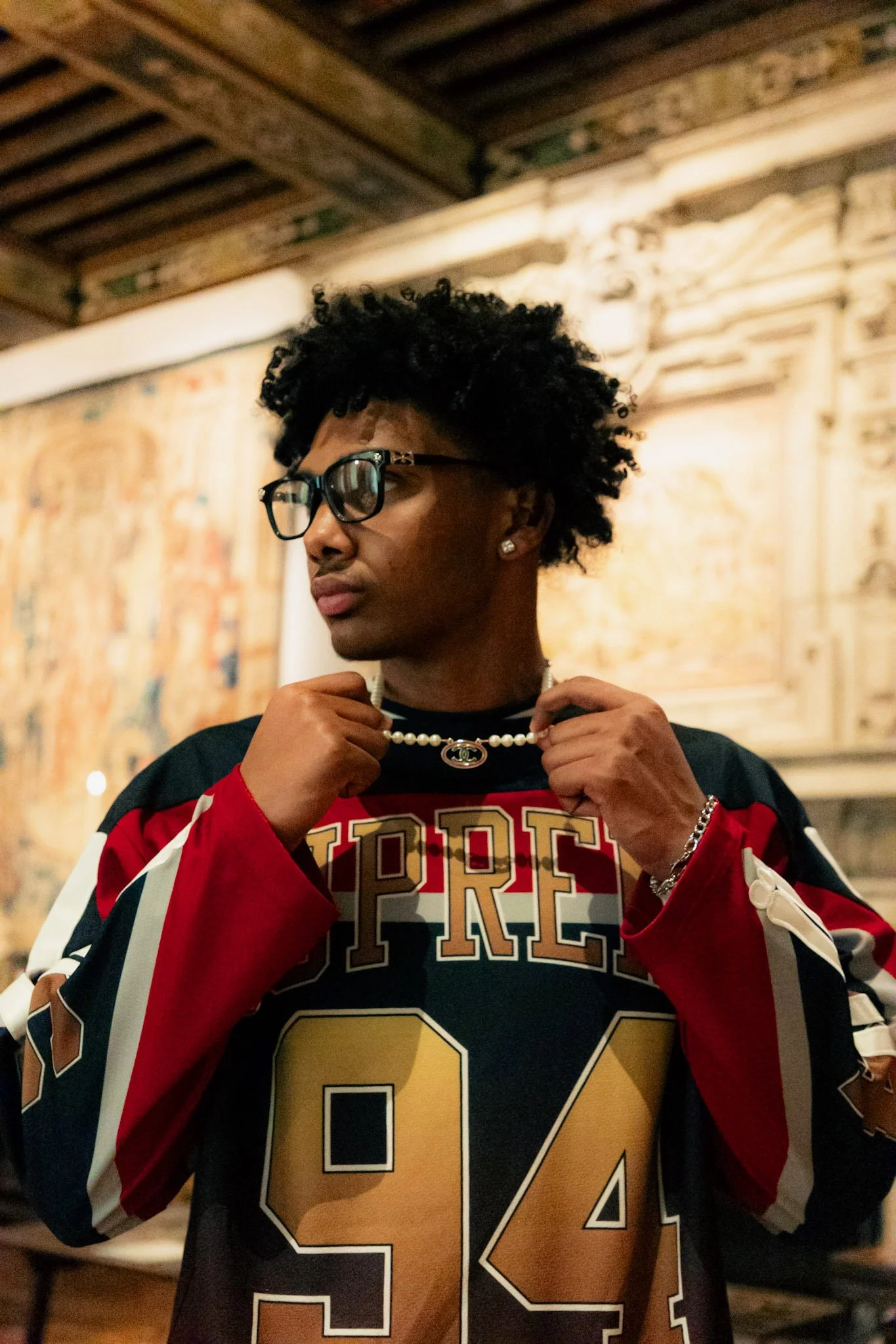 A young person wearing a black, red, and white sports jersey with the number 94, glasses, and jewelry, holding a pearl necklace with a designer logo in front of a textured wall with artwork.