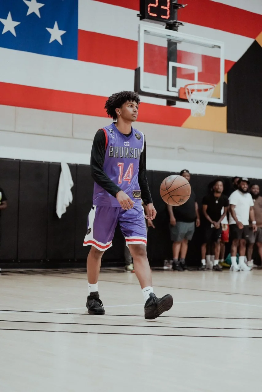 A young male basketball player wearing a purple and black jersey with the number 14, standing on an indoor basketball court with an American flag mural in the background. He is dribbling a basketball, and a scoreboard is visible above him.